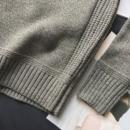 Close-up of a gray textured sweater with ribbed cuffs and hem on a white surface.