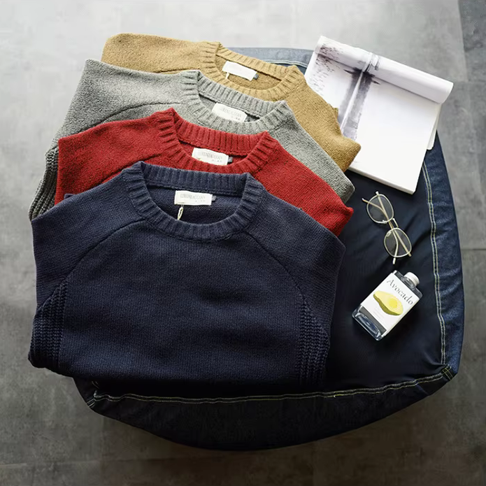 Stack of folded sweaters with a small bottle and sunglasses on a gray surface