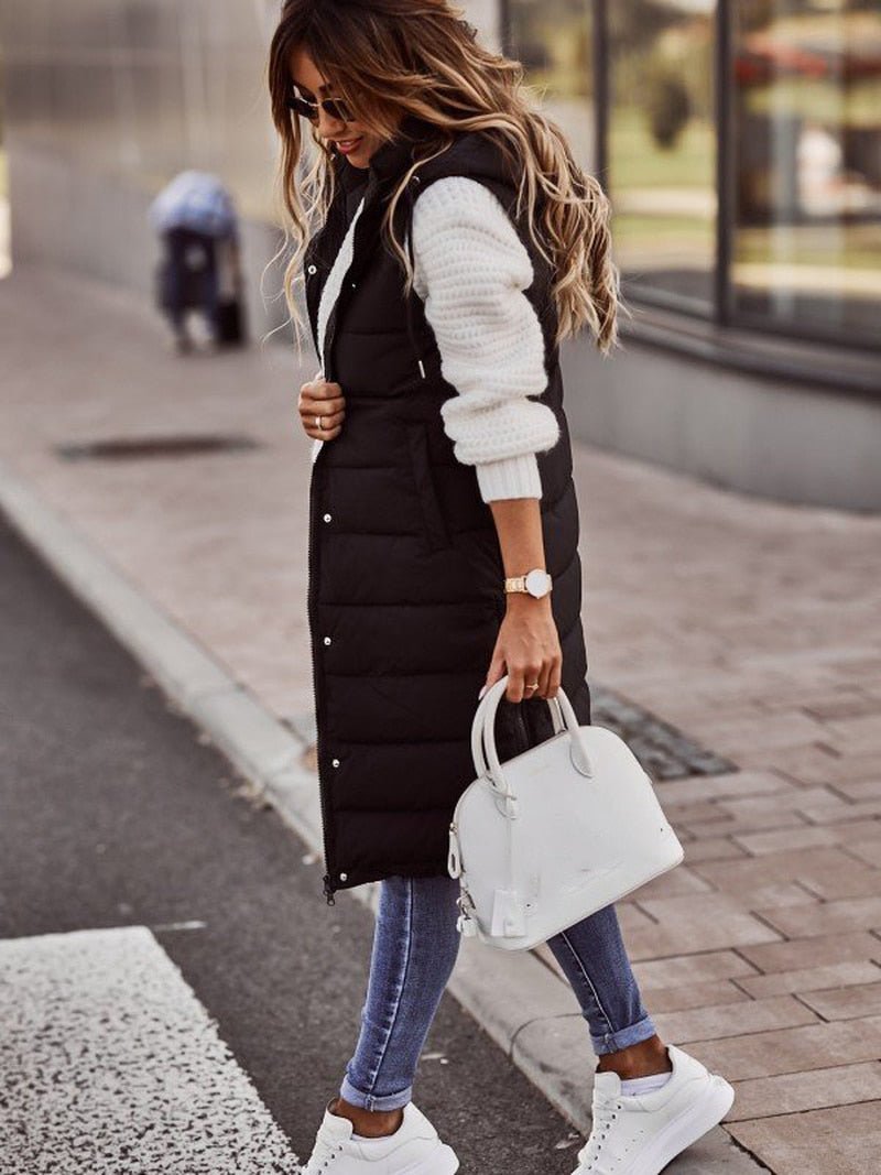 Woman wearing a black puffer vest, white sweater, blue jeans, and white sneakers on a city street.
