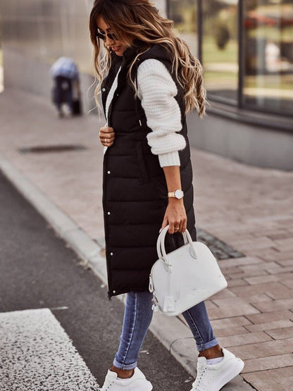 Woman wearing a black puffer vest, white sweater, blue jeans, and white sneakers on a city street.