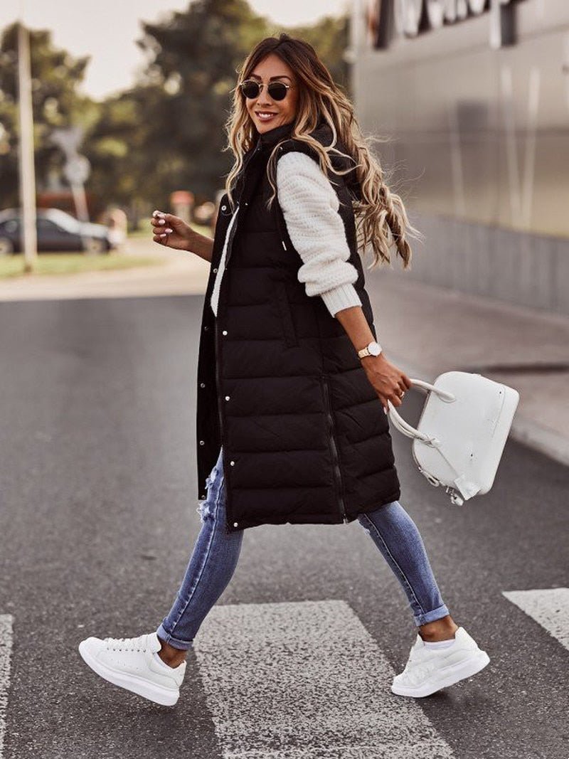 Woman walking outdoors wearing a black puffer vest, white sweater, blue jeans, and white sneakers.