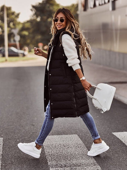 Woman walking outdoors wearing a black puffer vest, white sweater, blue jeans, and white sneakers.