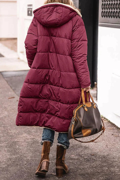 Person wearing a long burgundy puffer coat with a hood, holding a brown handbag on a sidewalk.