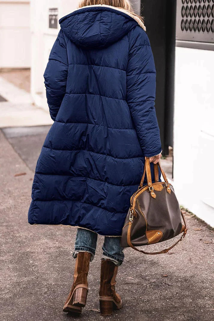 Person wearing a long blue puffer coat holding a brown handbag on a street.