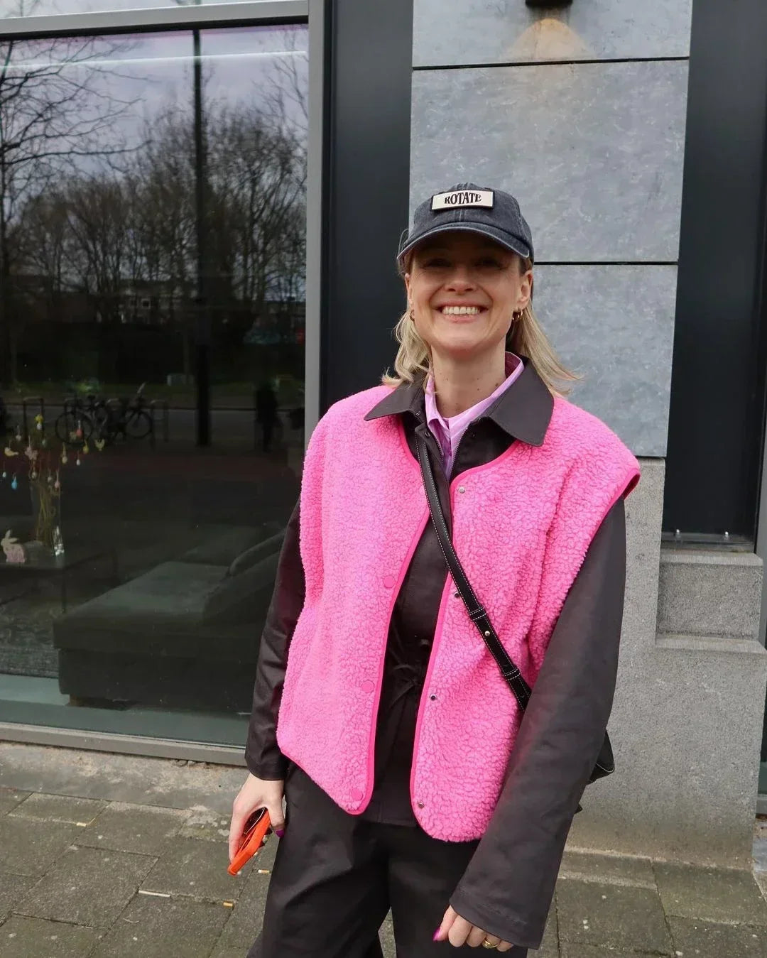 Person wearing a pink vest and black cap standing outside a building.