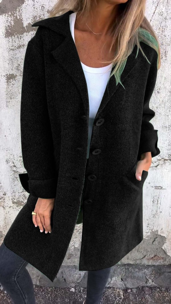 Person wearing a black coat with a white shirt underneath against a textured wall.