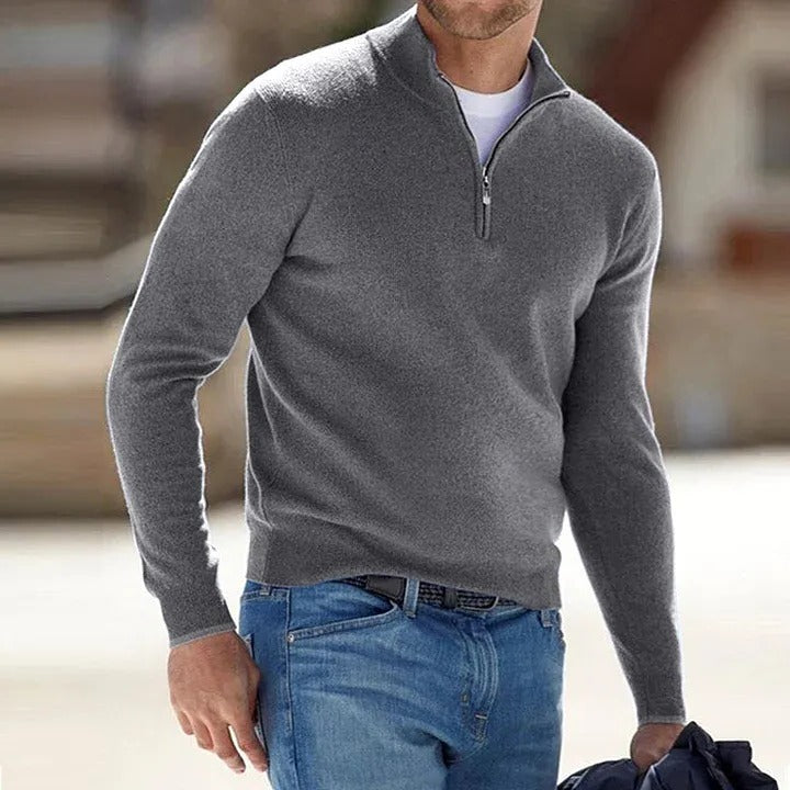 Man wearing a gray half-zip sweater and blue jeans with a blurred background