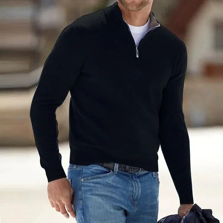 Man wearing a black quarter-zip sweater and blue jeans indoors.