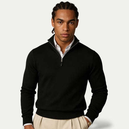 Man wearing a black quarter-zip sweater over a white shirt with beige pants on a light gray background