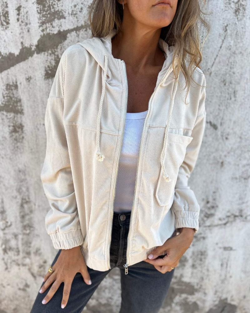 Woman wearing a beige jacket over a white top against a textured wall.